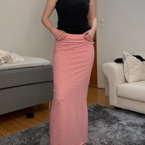 Rosa randig kjol - Säljer en super gullig rosa och vitrandig långkjol med en tight passform. Kjolen är perfekt för sommardagar och har en enkel men stilren design. Den är gjord i ett mjukt och bekvämt material som gör den skön att bära hela dagen. Materialet är stretchigt så det passar flera storlekar. Jag har brukar ha storlek 36/38 i kläder.