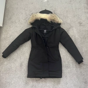 Svart parkajacka från Canada Goose - Säljer en svart parkajacka från Canada Goose med avtagbar päls på huvan. Jackan har en klassisk design med lång ärm och dragkedja framtill. Perfekt för kalla vinterdagar.