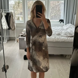 Mönstrad omlottklänning från Modes Stockholm - Snygg omlottklänning från Modes Stockholm i ett unikt mönster med jordnära toner. Klänningen har en v-ringad hals och trekvartsärmar, vilket ger en elegant touch. Perfekt för att sticka ut med stil! Vet ej storleken men uppskattar till S. Nypris är 1600kr