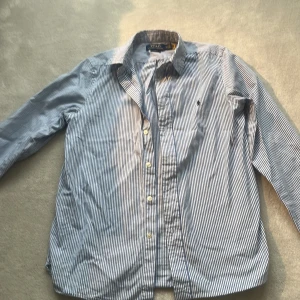 Randig skjorta från Polo Ralph Lauren - Snygg blå- och vitrandig skjorta från Polo Ralph Lauren i storlek L. Skjortan har långa ärmar och klassiska knappar framtill. Den ikoniska loggan är broderad på bröstet. Perfekt för en stilren look! Är i storlek 18 barn men passar xs