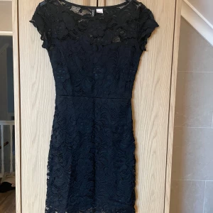 Svart spetsklänning från H&M - Svart spetsklänning från H&M med korta ärmar och rund hals. Klänningen har ett vackert blommönster i spetsen och en figurnära passform. Klänningen är i mycket bra skick!🥰