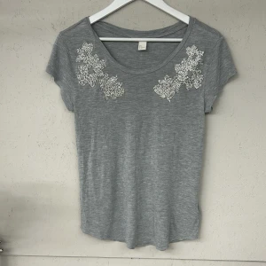 Grå t-shirt med spetsdetaljer från H&M - Säljer en stilren grå t-shirt från H&M med vackra spetsdetaljer på axlarna. T-shirten är kortärmad och tillverkad i mjukt material för en bekväm passform. Perfekt för en avslappnad look.