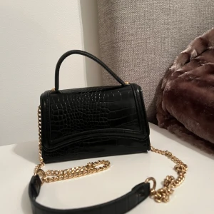 Svart axelväska med kedja - Snygg svart axelväska i syntet med krokodilpräglat mönster och guldfärgad kedja. Väskan har ett handtag och axelrem kedja. Som helt ny.