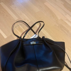 Helt ny Svart axelväska Gina tricot - Snygg svart väska  med stilren design. Väskan har dubbla handtag och en elegant låsdetalj framtill. Perfekt för att bära med sig det nödvändigaste med stil. Aldrig använd!