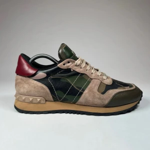 Valentino Rockrunner sneakers i mocka och camouflage - Snygga Valentino Rockrunner sneakers med en cool camouflagedesign. Skorna har en mix av mocka och syntetmaterial i beige, grön och röd. De har snörning och en unik nitat detalj på hälen. Perfekta för att ge din stil en edgy touch.