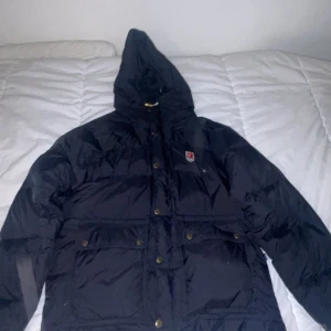 Fjällräven jacka - Säljer en svart dunjacka från Fjällräven med huva och praktiska fickor. Jackan har en dragkedja och knappar framtill samt justerbara snören i huvan. Perfekt för kyliga dagar. självaste jackan är i fint skick och det är en väldigt skön jacka att använda , Kan mötas upp nära malmö 👍