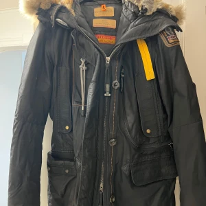 Svart vinterjacka från Parajumpers - Säljer en svart vinterjacka från Parajumpers med pälsfodrad huva och flera praktiska fickor. Jackan har en dragkedja och knappar framtill samt en gul detalj vid kragen. Perfekt för kalla dagar.