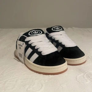 Svarta Adidas Campus sneakers - Snygga Adidas Campus sneakers i svart. Skorna har en vit sula och bruna yttersulor. Tillverkade i mocka med snörning och Adidas logga på plösen och hälen. Perfekta för en stilren look. Alla accessoarer är inkluderade!
