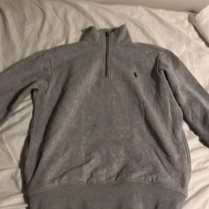 Grå tröja från Polo Ralph Lauren - Säljer en stilren grå tröja från Polo Ralph Lauren med halv dragkedja och broderad logotyp på bröstet. Perfekt för en avslappnad look. Tröjan har långa ärmar och är tillverkad i ett mjukt material. Tröjan är sparsamt använd och priset är ej hugget i sten