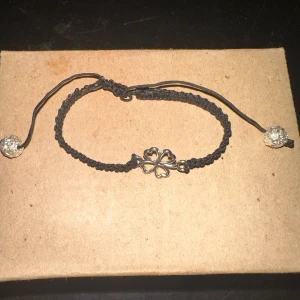 Svart armband med fyrklöver - Säljer ett snyggt svart armband med en fyrklöver i mitten. Armbandet är justerbart och har glittriga pärlor i ändarna. Perfekt för att ge lite tur och stil till din outfit!
