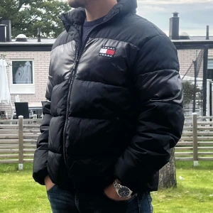 Svart pufferjacka från Tommy Jeans - Säljer en snygg svart pufferjacka från Tommy Jeans. Jackan har en dragkedja framtill och är perfekt för kyligare dagar. Den är i bra skick och har en klassisk design med märkets logga på bröstet. Perfekt för både höst och vinter!