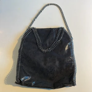 Svart axelväska med kedjedetaljer - Säljer en  svart axelväska med kedjedetaljer runt kanterna och som axelrem.. Den är rymlig och passar både till vardags och fest. Väskan är ny och oanvänd.