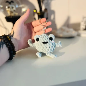 Söt nyckelring med virkad figur - Den här nyckelringen har en supergullig virkad figur i ljusgrått med stora svarta ögon och ett glatt leende. Perfekt att hänga på väskan eller nycklarna för en extra touch av personlighet!
