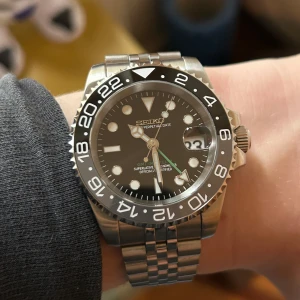 Seiko mod GMT master ”Bruce Wayne” - Snygg Seiko mod klocka med svart urtavla och lysande markeringar. Den har en silverfärgad boett och armband i rostfritt stål, bredden på boet är 40mm. Klockan visar datum och har en vridbar bezel med 24-timmarsmarkeringar. NH34 urverk med GMT funktion. Dm vid frågor eller prisförslag 😁📩