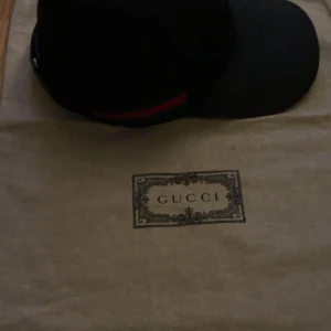 Svart keps från Gucci - Snygg svart keps från Gucci med det klassiska GG-mönstret. Kepsen har en röd och grön randdetalj på sidan och är perfekt för en stilren look. Och äkta skriv kan skicka mer haft i 1 år storlek xs