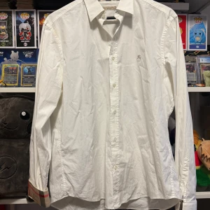 Vit skjorta från Burberry - Snygg vit skjorta från Burberry i bomull med klassisk krage och knappar. Den har långa ärmar och ett diskret broderat märke på bröstet. Insidan av kragen har ett stilrent rutigt mönster. Den behöver en tvätt för att se ny ut annars finns det inga fel.