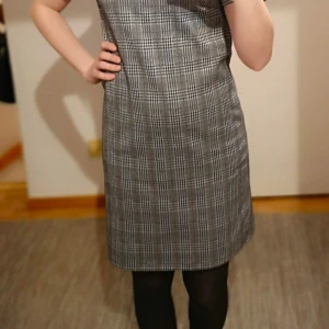 Grårutig klänning från Vila - Snygg rutig klänning från Vila i grå, vit och svart. Den har en klassisk rak passform med korta ärmar och en dragkedja i ryggen. Storlek XS. Perfekt för att användas till högtiderna ❤️