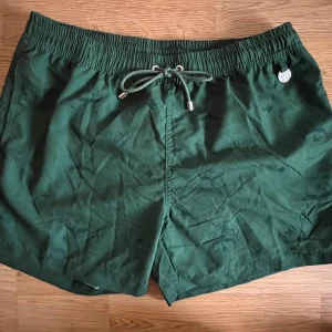 Gröna shorts (Studio STHLM) - Snygga gröna badshorts från Studio STHLM med snörning i midjan och ett diskret mönster. Perfekta för en avslappnad stil. De har en praktisk bakficka med dragkedja. Oanvända!  Originalpris: 499kr (Finns katt i hushållet)