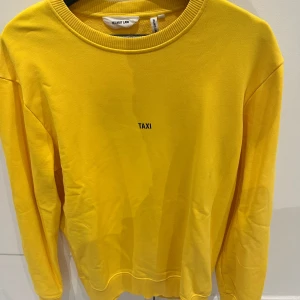 Gul tröja från Helmut Lang - Säljer en gul tröja från Helmut Lang med texten 'TAXI' på framsidan och ett tryck på baksidan. Tröjan har långa ärmar och en rund halsringning. Perfekt för dig som vill sticka ut med en unik design!