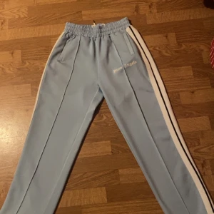 Ljusblå träningsbyxor från Palm Angels - Snygga ljusblå träningsbyxor från Palm Angels med vita sidoremsor och elastisk midja. Byxorna har en avslappnad passform och är perfekta för en sportig look. Märkesloggan är tryckt på framsidan.