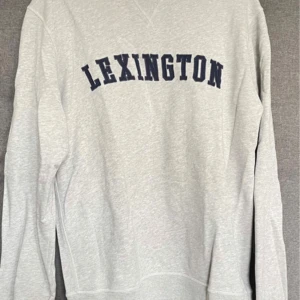 Grå sweatshirt från Lexington - Säljer en stilren grå sweatshirt från Lexington med mörkblå text på bröstet. Tröjan är tillverkad i 100% bomull och har långa ärmar. Perfekt för en avslappnad stil.