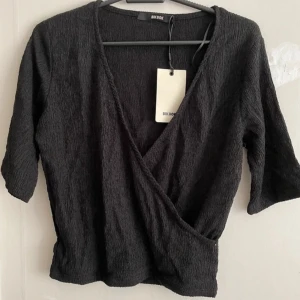 Svart omlottopp från Bik Bok - Snygg svart omlottopp från Bik Bok med trekvartsärmar. Toppen har en v-ringad design och är perfekt för en stilren look. Passar bra till både jeans och kjol.