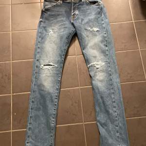 Säljer ett par blå jeans med coola slitningar på låren och knäna. Perfekta för en avslappnad stil. De har en klassisk femficksdesign och knappgylf.