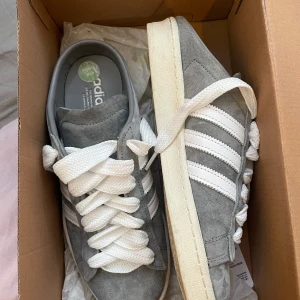 Grå Adidas sneakers - Säljer ett par stilrena grå Adidas sneakers i mocka med vita ränder och snörning. Perfekta för en avslappnad look. Endast använt 4 gånger så nästan nyskick. Skriv om ni har frågor!