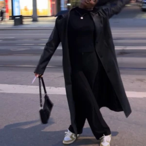 Svart trenchcoat från PrettyLittleThing - Snygg svart trenchcoat från PrettyLittleThing i syntetmaterial. Perfekt för en stilren look med långa ärmar och klassisk design. Passar utmärkt för både höst och vår.