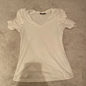 Vit topp från KappAhl - Säljer en söt vit topp från KappAhl i storlek XS. Toppen har korta ärmar med rynkade detaljer och en v-ringad hals. Perfekt för en stilren look.