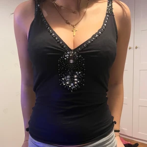 Svart linne från More & More - Snyggt svart linne från More & More med paljettdetaljer vid halsringningen och framtill. Linnet har en v-ringad design och ärmlös stil, perfekt för en kväll ute. Passar bra till både jeans och kjol. Insydd i midjan men inget som syns