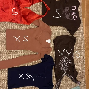 Fem olika toppar i varierande stilar och storlekar - Samling av fem toppar: en röd satin halterneck i storlek S = 150, en brun  D&G kortärmad topp i storlek S= 170, en brun ribbad långärmad topp i storlek XS= 90, en svart paljettprydd ärmlös topp i storlek XXS =120, och en mörkblå  topp i storlek XS = 150.  PRISER VARIERAR KÖP ALLA FÖR 400