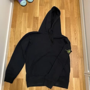 Svart hoodie från Stone Island - Säljer en stilren svart hoodie från Stone Island med deras ikoniska märke på ärmen. Tröjan har en klassisk huva med dragsko och långa ärmar. Perfekt för en avslappnad look.