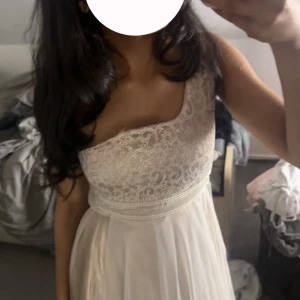 Vit spetsklänning med knytband - Elegant vit klänning med spetsdetaljer på överdelen och ett knytband i midjan. Klänningen har en ärmlös design och en lång, flödande kjol. Perfekt för somriga tillfällen.