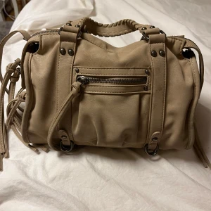 Beige axelväska med dragkedja - Snygg beige axelväska med flera dragkedjor och flätade handtag. Väskan har dekorativa knytband och metallnitar som ger en cool look. Perfekt för att bära dina dagliga nödvändigheter med stil.