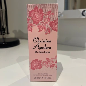 Christina Aguilera Definition Eau de Parfum - Elegant parfymflaska med en feminin design, dekorerad med rosa blommor och en svart rosett. Doften är en Eau de Parfum och kommer i en 30 ml flaska. Helt ny!