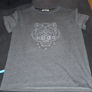Grå t-shirt med tigertryck från Kenzo - Grå t-shirt från Kenzo med det ikoniska tigertrycket och texten 'Kenzo Paris' på framsidan. Perfekt för en stilren och modern look.