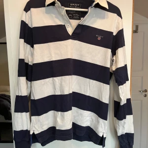 Randig Rugbytröja från GANT - Säljer en klassisk randig pikétröja från GANT i marinblått och vitt. Tröjan har långa ärmar och en stilren krage med knappar. Perfekt för en avslappnad men stilfull look.