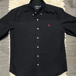 Polo Ralph Lauren Oxford Skjorta Navy - Snygg svart skjorta från Polo Ralph Lauren med klassisk krage och knappar framtill. Den har en broderad röd logotyp på bröstet som ger en stilren touch. Perfekt för en elegant look.