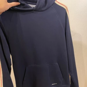 Mörkblå hoodie från GANT - Säljer en snygg mörkblå hoodie från GANT med vita och röda ränder längs ärmarna. Tröjan har en klassisk känguruficka och en bekväm huva. Perfekt för en avslappnad stil.