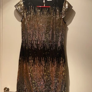 Glittrig klänning från KappAhl st 170 (xs/s) - Snygg glittrig klänning från KappAhl med paljetter i guld,silver och metalliska skimrande färger. Den har korta ärmar och en elegant svart baksida. Perfekt för en festlig kväll! Klänning har en under klänning så inget som ligger emot och skaver. St 170 men passar bra på xs/s! Super fin till NY ÅR! 