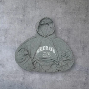  hoodie från Reebok 90’s - Säljer en grå hoodie från Reebok med stor logga framtill. Den har en klassisk design med huva och ribbade muddar vid ärmslut och nederkant. Perfekt för en avslappnad stil.