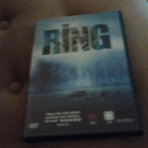 The Ring DVD - Säljer en DVD av skräckfilmen 'The Ring'. Omslaget är mörkt och mystiskt med en suddig bild av en brunn och titeln i stora bokstäver. Perfekt för filmkvällar med vänner som gillar att bli skrämda.