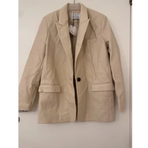 Beige kavaj med knappar - Snygg beige kavaj med klassisk design och svarta knappar. Perfekt för en stilren look. Långärmad med enkel knäppning framtill och fickdetaljer. Oanvänd (prislapp kvar). 