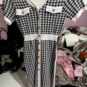 Svartvit klänning med knappar - Snygg svartvit klänning med hundtandsmönster och korta ärmar. Klänningen har en v-ringad design med dekorativa knappar framtill och två fickdetaljer på bröstet. Perfekt för en chic look!