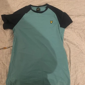 Turkos och marinblå t-shirt från Lyle & Scott - Snygg t-shirt från Lyle & Scott i turkos med marinblå ärmar och krage. Den har en liten broderad logga på bröstet. Perfekt för en avslappnad stil. Och har ett litet hål på baksidan