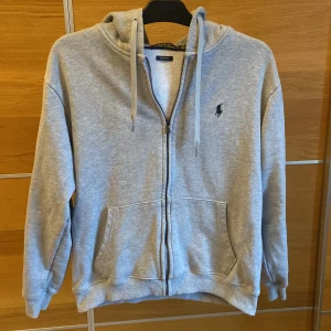 Grå hoodie från Polo Ralph Lauren - Säljer en stilren grå hoodie från Polo Ralph Lauren med dragkedja och justerbar huva. Tröjan har en broderad logga på bröstet och praktiska fickor framtill. Perfekt för en avslappnad look. Den är en M fast i BARN storlek så den är liten som ifall man har herr kläder så är det en S skulle man kunna säga! Hör av er ifall ni är intresserade!🙌