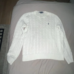 Vit stickad tröja från Ralph Lauren - Säljer en klassisk vit stickad tröja från Ralph Lauren med kabelstickat mönster och broderad logotyp på bröstet. Tröjan har långa ärmar och ribbade muddar vid ärmslut och nederkant. Perfekt för kyliga dagar!