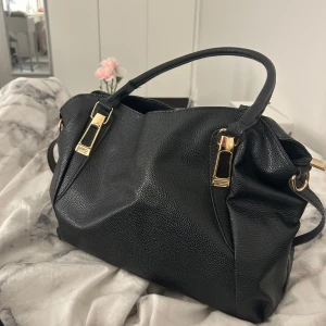 Svart handväska med gulddetaljer - Snygg svart handväska i skinn med eleganta gulddetaljer. Väskan har en praktisk dragkedja och flera fack inuti för enkel organisering. Perfekt för skolan och till vardag.