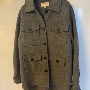 Grå ulljacka från Michael Kors - Snygg grå ulljacka från Michael Kors med knappar framtill och fyra praktiska fickor. Jackan har en klassisk krage och långa ärmar, perfekt för kyligare dagar.